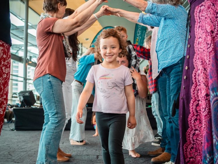 Image of A young girl smiles whilst walking barefoot through a tunnel formed by adults holding hands inside a bright, modern room. Other people stand in the background, creating a lively, joyful atmosphere.
