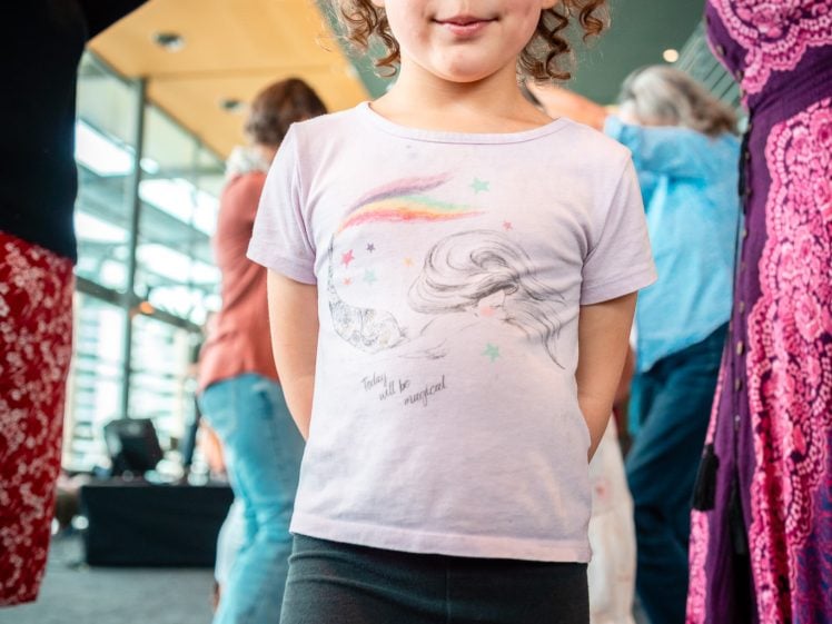 Image of A young child with curly hair, wearing a unicorn T-shirt and black trousers, stands indoors smiling at the camera whilst adults dance in the background.
