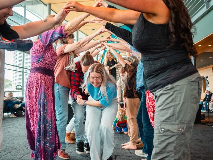 Image of A smiling woman in light blue and white clothes bends forward, moving through a lively human tunnel formed by people joining raised hands indoors, with others cheering and watching.