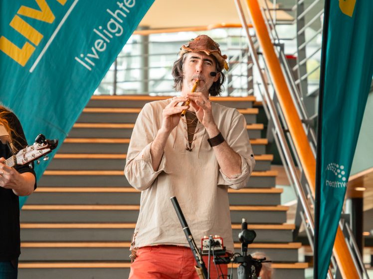Image of A musician in a hat and red trousers plays a wooden flute on stage in front of stairs, with Live Lighter banners and microphones visible beside him at an indoor event.