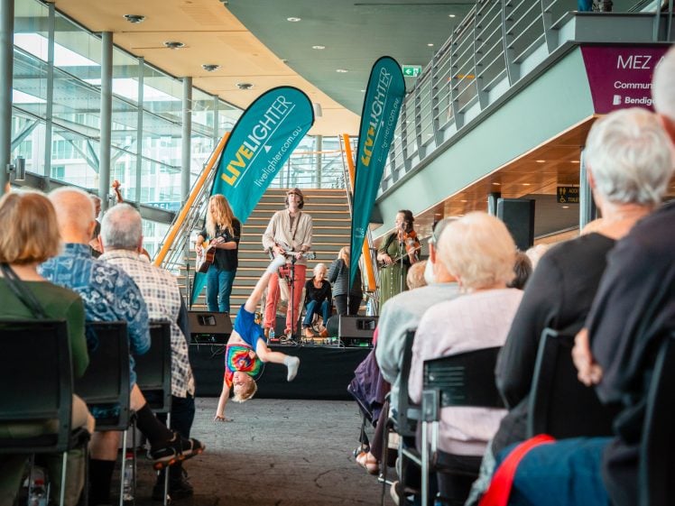 Image of A breakdancer performs a handstand on a carpeted aisle as a live band plays on stage to an audience seated in a modern indoor venue with glass walls and balcony.