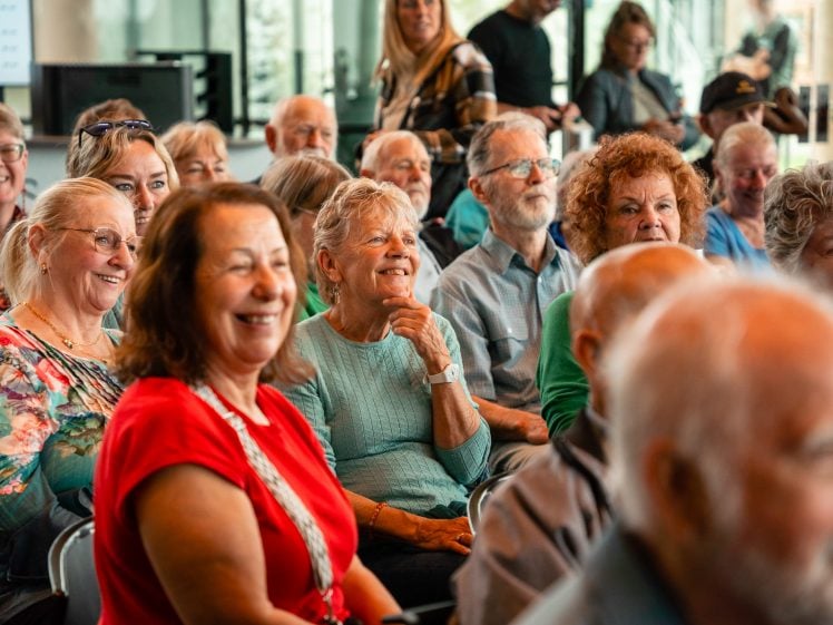 Image of A group of older adults sit closely together, smiling and laughing at an indoor event, creating a warm and cheerful atmosphere. The background shows more people and large windows letting in natural light.
