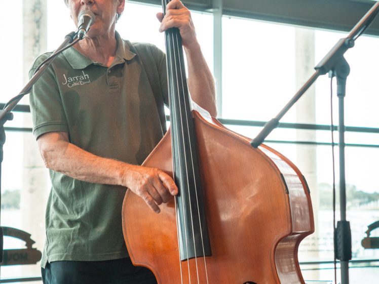 Image of An older man in a green shirt plays a double bass and sings into a microphone in a bright, modern indoor space. People sit in the background, and large windows let in natural light.