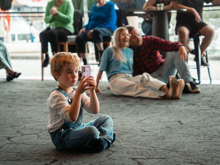 Image of A young child in dungarees sits cross-legged on a carpeted floor, holding up a mobile. In the background, a smiling couple poses for the camera, with other adults seated and watching nearby.