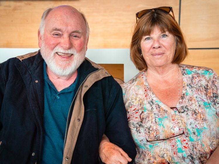 Image of An older man with a white beard and a woman with shoulder-length brown hair sit closely together, smiling at the camera. The man wears a dark jacket; the woman wears a colourful blouse and sunglasses on her head.