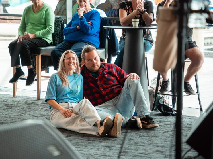 Image of A man and woman sit on the floor smiling, leaning against each other, while four older adults watch from chairs and a high table in a brightly lit indoor space with large windows.