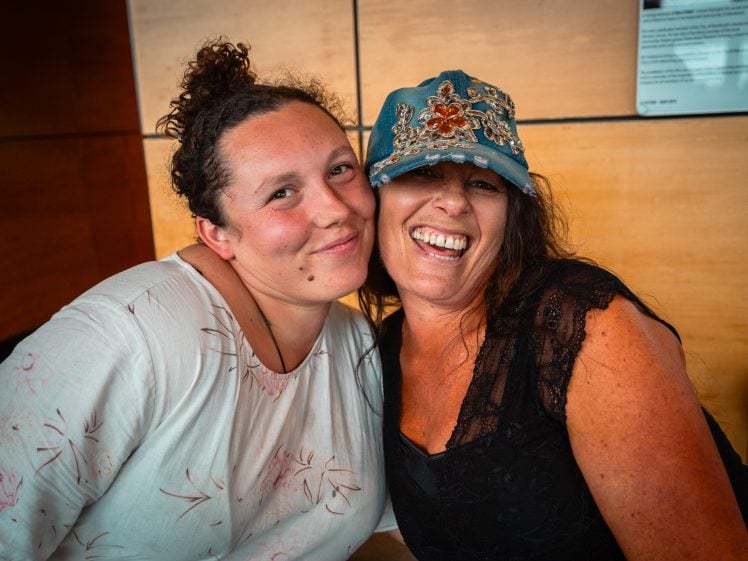 Image of Two women smile and pose closely together indoors; one wears a white patterned shirt, and the other wears a decorated blue cap and a black lace top. They appear happy and relaxed in a warmly lit setting.