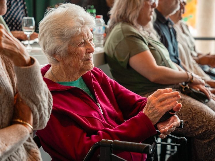 Image of An elderly woman with white hair, wearing a red jacket, sits in a wheelchair and claps her hands. She is surrounded by other seated people in a well-lit indoor setting.