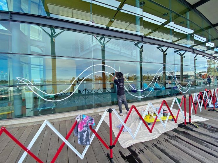 Image of An artist in a hat paints a large white wave pattern on the glass window of a modern building. Art supplies and colourful fabric are on the ground, and a red barrier lines the wooden decking outside.