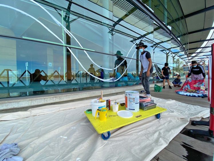 Image of Artists wearing masks work on painting a large glass window mural outside, with painting supplies on a yellow trolley in the foreground and dust sheets covering the ground.