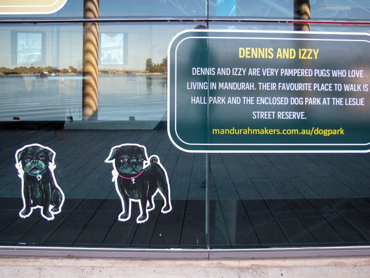 Image of A window displays two cartoon pugs, one sitting and one standing. Next to them is a sign about Dennis and Izzy, pampered pugs in Mandurah, with a website link for more info about the local dog park.