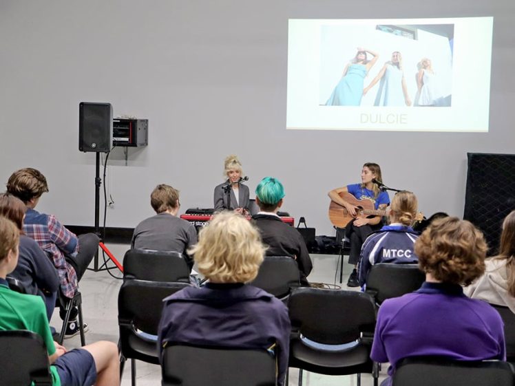 Image of Two musicians perform for an audience in a classroom. One plays keyboard and sings, the other plays guitar. A screen behind them displays a photo of three women and the word “DULCIE.” Audience members face the performers.