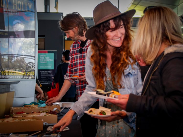 Image of Two women smiling and serving themselves food from a buffet table, with another person in the background; indoor setting with posters and windows visible.