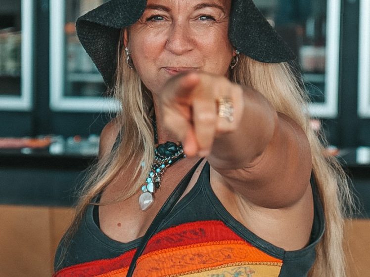 Image of A woman with long blonde hair wearing a black hat and a rainbow-striped vest points directly at the camera, smiling confidently. Shelves with bottles are visible in the background.