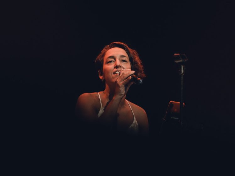 Image of A woman with short brown hair sings into a microphone on stage, illuminated by a spotlight against a dark background.