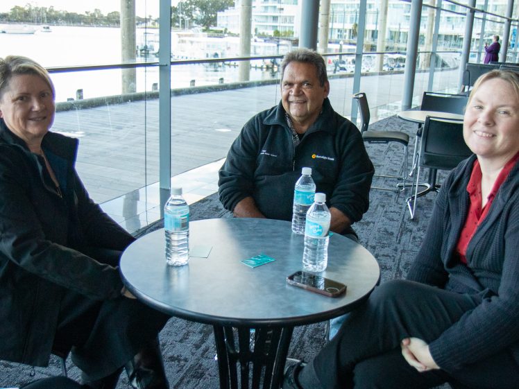 Image of Three people sit around a small round table indoors, each with a bottle of water. Large windows behind them show a waterfront view with boats and modern buildings. All three are smiling at the camera.