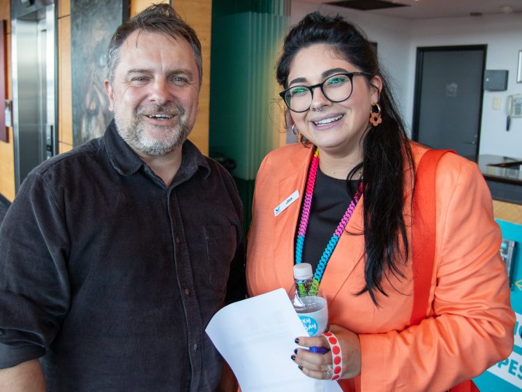 Image of A man in a dark shirt stands smiling next to a woman in an orange jacket and glasses, holding a water bottle and papers. They are indoors, appearing happy and friendly.