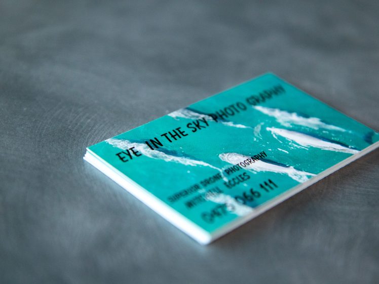 Image of A business card with an aerial photo of whales swimming in blue water, reading EYE IN THE SKY PHOTOGRAPHY along with contact information, placed on a grey textured surface.