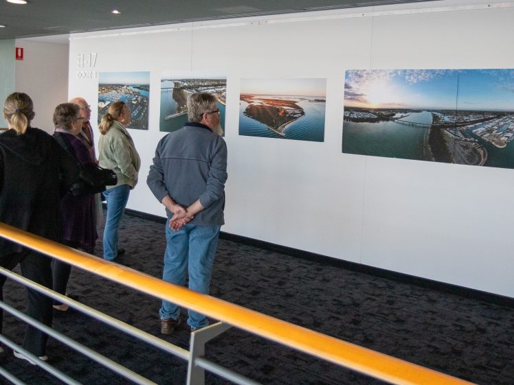 Image of People stand in a corridor, viewing several large aerial landscape photographs mounted on a white wall. The scene is indoors with natural light coming from the left.