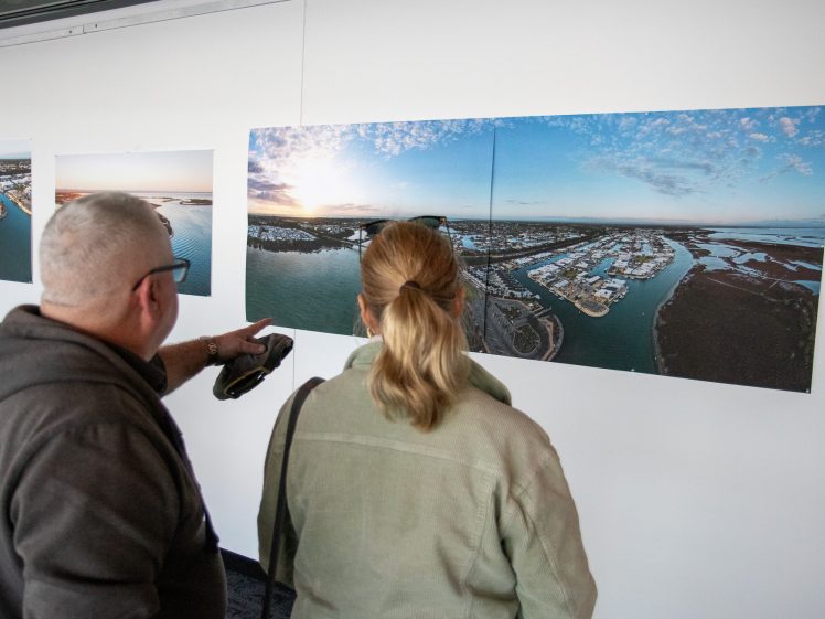 Image of Two people stand indoors looking at a large aerial photograph of a coastal area with water, land, and buildings, displayed on a white wall. The man points towards the image whilst the woman looks at it.