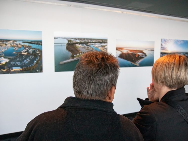 Image of Two people view a gallery wall displaying large aerial photographs of coastal landscapes. They stand with their backs to the camera, closely observing the images together.