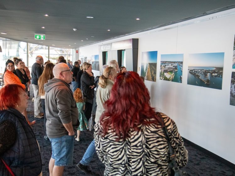 Image of A group of people stand in a gallery space, viewing large aerial landscape photographs displayed on a white wall. The crowd faces the artwork, engaged and observing the images.
