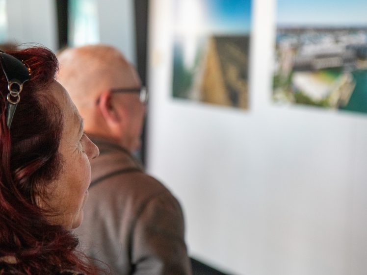 Image of Two people, seen from behind, closely observe framed photographs or artwork displayed on a white wall in a gallery or exhibition setting. The focus is on their heads and shoulders.