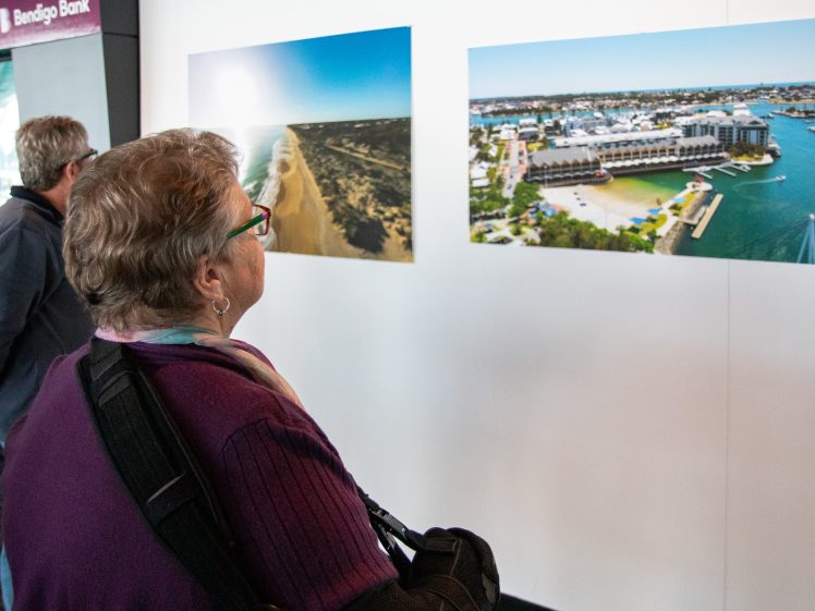 Image of Two people stand indoors, closely observing two large aerial photographs of a beach and a marina displayed on a white wall. One person wears glasses and a purple jumper.