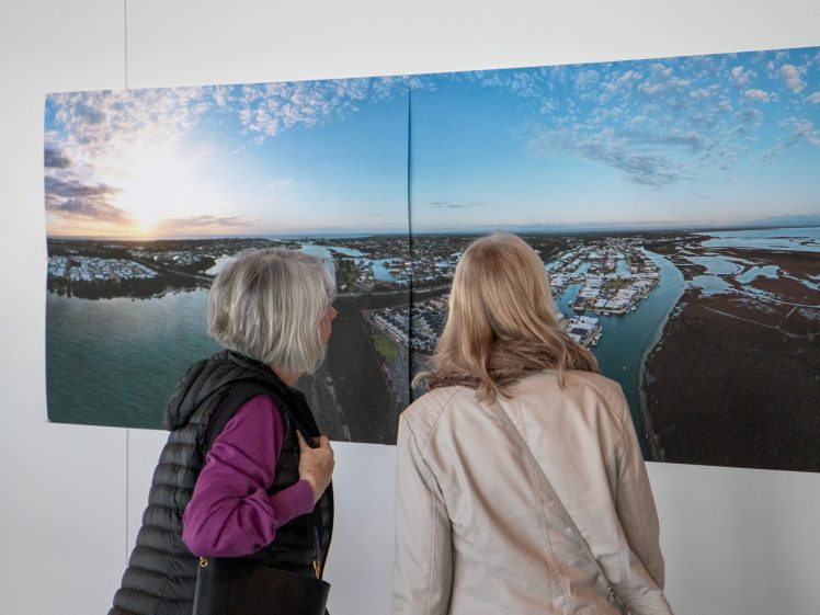 Image of Two women stand closely together, facing and observing a large panoramic photograph of a coastal landscape displayed on a white wall in an art gallery.