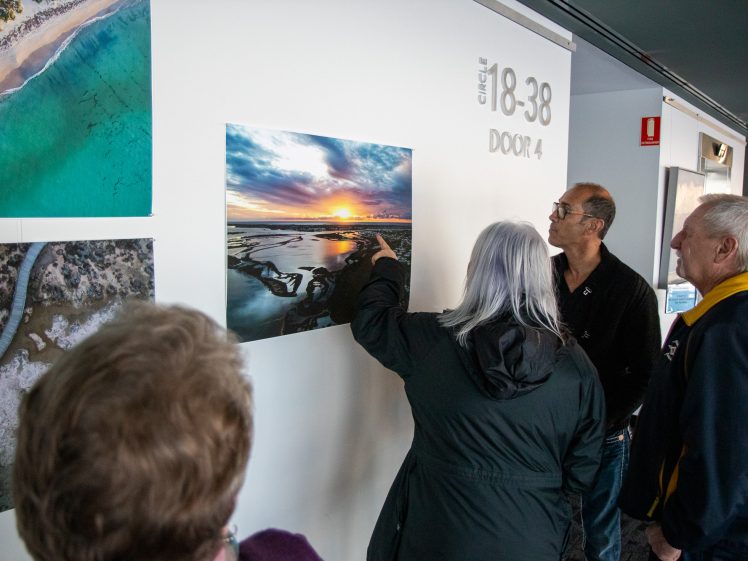 Image of Four people stand in front of a wall displaying aerial landscape photos. One person points at a photo of a sunset over water whilst the others look on and discuss the image.