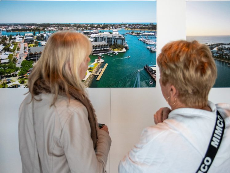 Image of Two women with fair hair and light-coloured jackets stand indoors, closely observing a large aerial photo of a marina with buildings and boats on display. Another landscape photo is visible next to it.