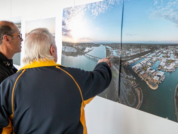 Image of Two men stand indoors, closely examining and discussing a large aerial photograph of a coastal cityscape with rivers, houses, and a bridge, which is displayed on a white wall. One man is pointing at the image.