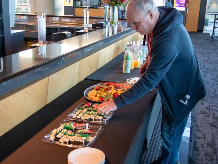 Image of A man in a hoodie selects sushi from a catering table with trays of sushi, fruit, and bottled drinks in a modern indoor setting.