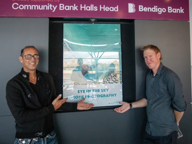 Image of Two men stand on either side of a window display poster for Eye in the Sky Drone Photography, with Community Bank Halls Head and Bendigo Bank signage visible above them.