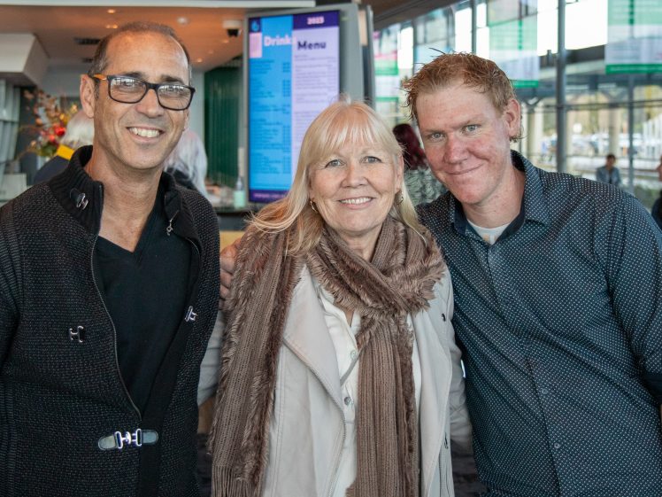 Image of Three people stand together, smiling for the camera indoors. The two men are on either side of a woman wearing a light coat and scarf. A digital drinks menu and windows are visible in the background.