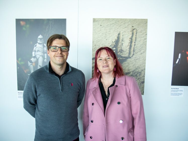 Image of Two people stand side by side, smiling in front of a white wall displaying photographs. The person on the left wears glasses and a grey jumper; the person on the right has red hair and wears a pink jacket.