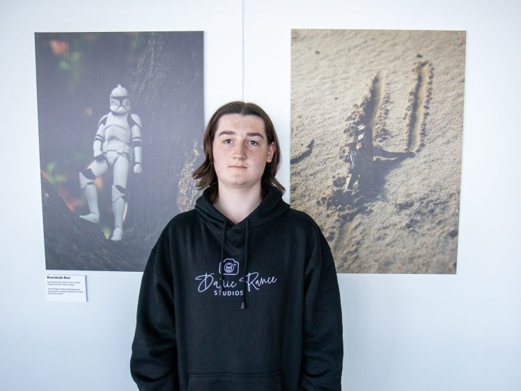 Image of A person with long brown hair wearing a black hoodie stands in front of two photos on a wall—one of a Star Wars clone trooper toy in a forest, and one of animal tracks in sand.