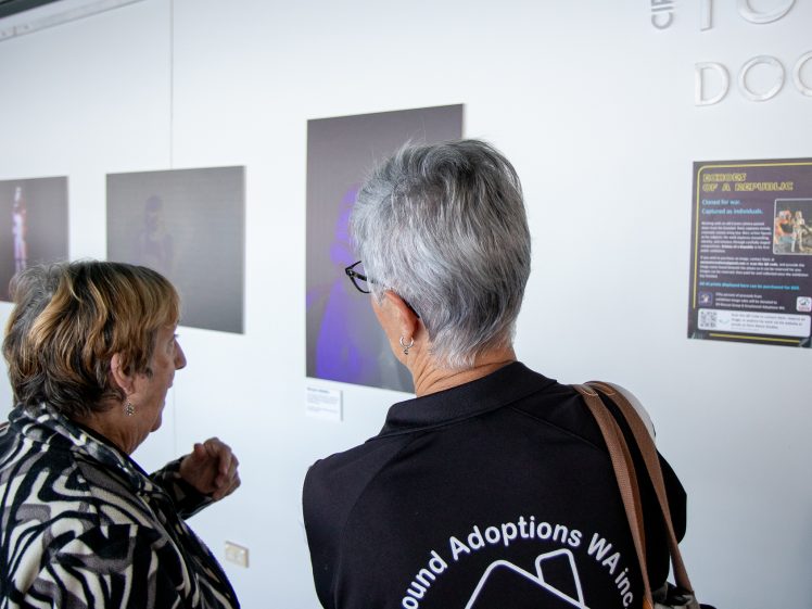 Image of Two women stand in front of framed artwork displayed on a wall, viewing and discussing the pieces. One woman wears a black shirt with a white logo on the back. A poster with text is visible to the right.