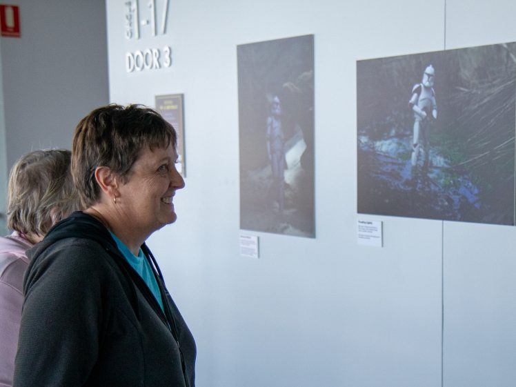 Image of Two women stand in front of a white wall, smiling as they look at two framed photos featuring Star Wars clone troopers in natural outdoor settings.