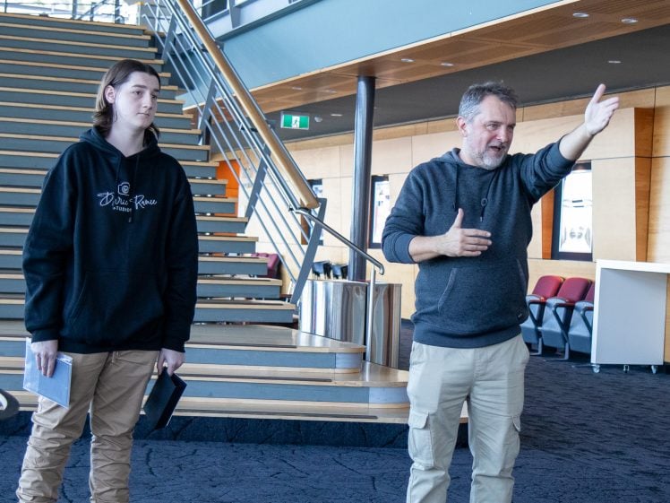 Image of Two people stand indoors near a staircase. One gestures with his arm extended and appears to be giving instructions or explaining something. The other holds a notebook and listens attentively. They are in a modern, well-lit building.