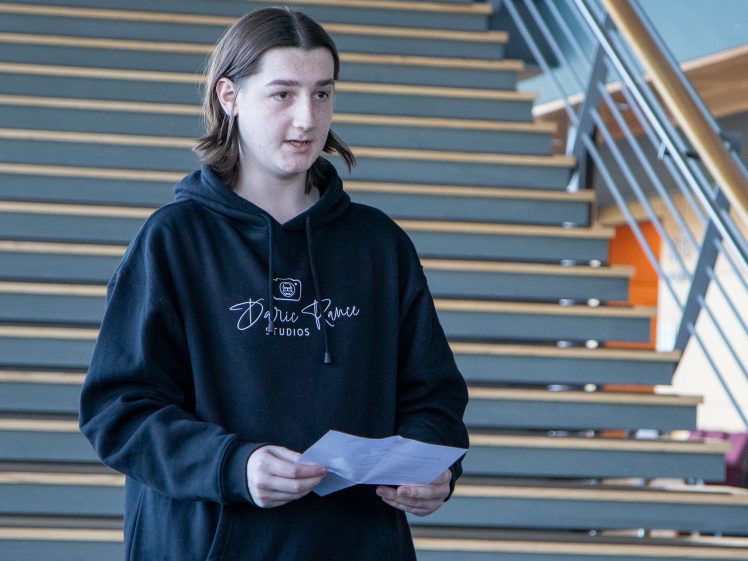 Image of A young person with long brown hair, wearing a black hoodie, stands indoors holding a sheet of paper in front of a large staircase.