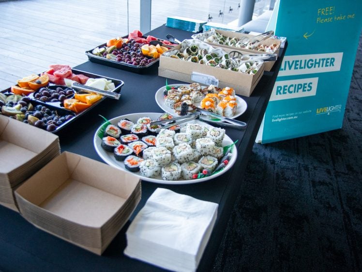 Image of A table with plates of sushi, fruit, wraps, and snacks, next to empty cardboard food boxes and serviettes. A blue sign reads Livelighter Recipes and Free! Please take one.