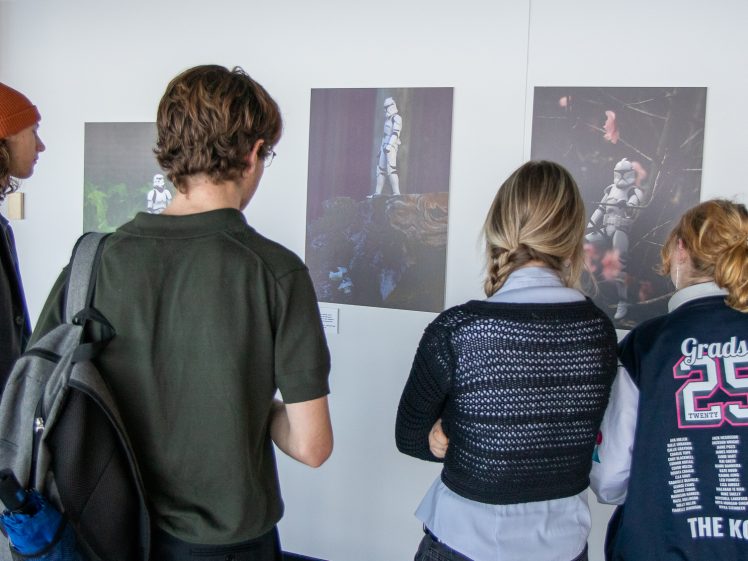 Image of Four people stand facing a wall, looking at photographs of stormtroopers on display. One person wears an orange beanie, another has a rucksack, and one wears a jacket with Grads 25 on the back.