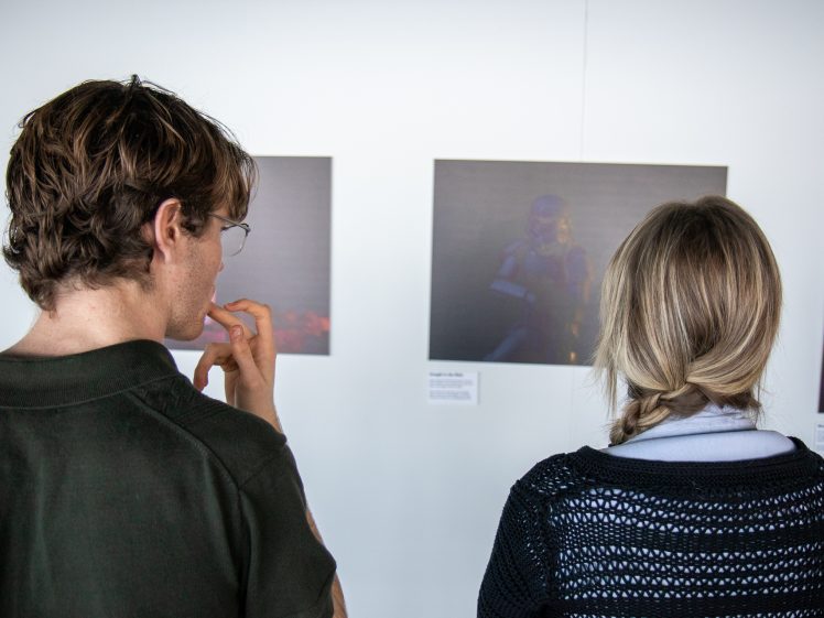 Image of Two people stand with their backs to the camera, viewing framed artwork displayed on a white wall in a gallery setting. One person has short brown hair, whilst the other has long blonde hair in a plait.