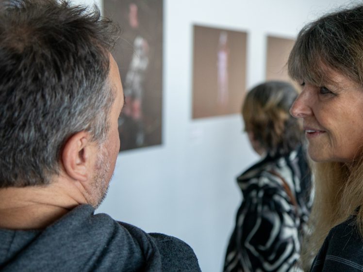 Image of Two people are conversing whilst viewing art in a gallery; another person stands ahead, looking at framed artworks displayed on a white wall.