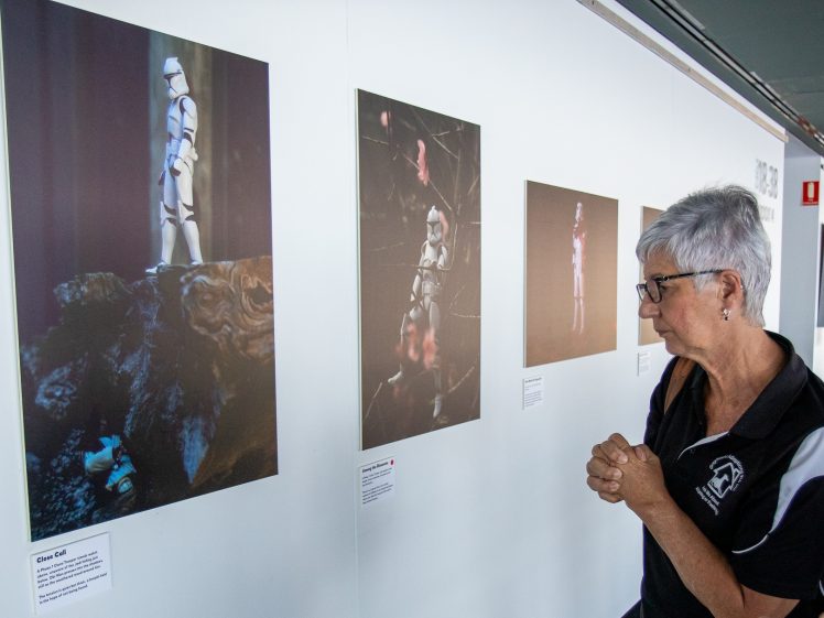 Image of An older woman with short grey hair and glasses looks closely at photos of Star Wars stormtrooper figurines displayed on a white gallery wall.