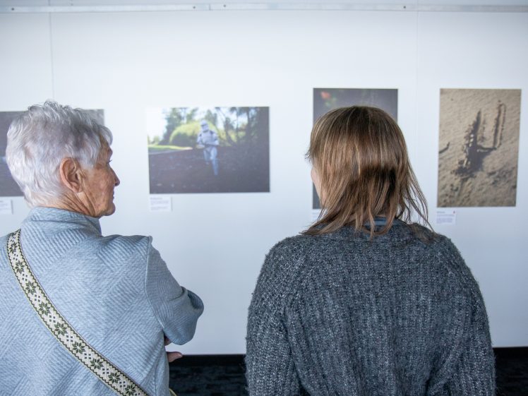Image of Two people with their backs to the camera view photographs displayed on a white gallery wall. One has short grey hair and the other has brown hair. The exhibited photos feature outdoor scenes.