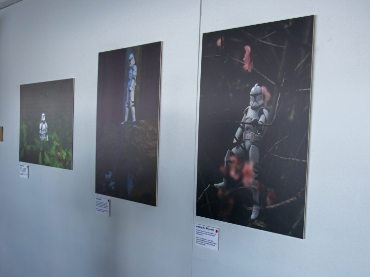 Image of Three framed photographs of toy stormtroopers from Star Wars are displayed on a white wall, each showing the figure in different outdoor settings with foliage and nature. Descriptive labels are beneath each artwork.