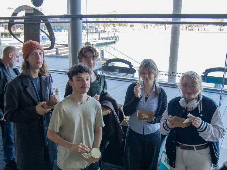 Image of Five young adults stand indoors near large windows, holding food containers and cups, as they pose for a photo. A waterfront view and dock are visible outside in the background.