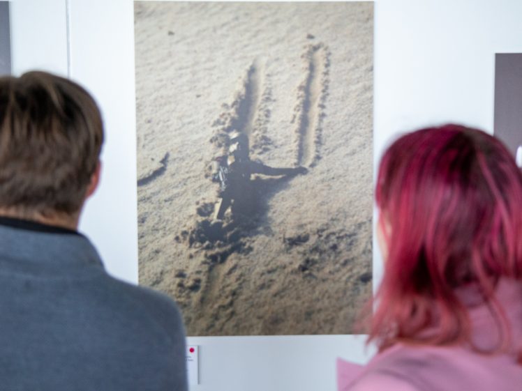 Image of Two people with their backs to the camera view a photograph of marks in the sand displayed on a white wall. The person on the left has short brown hair; the person on the right has shoulder-length pink hair.
